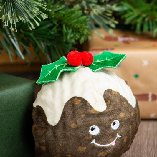 Plush Christmas pudding toy with a face and holly leaves in front of a decorated Christmas tree.