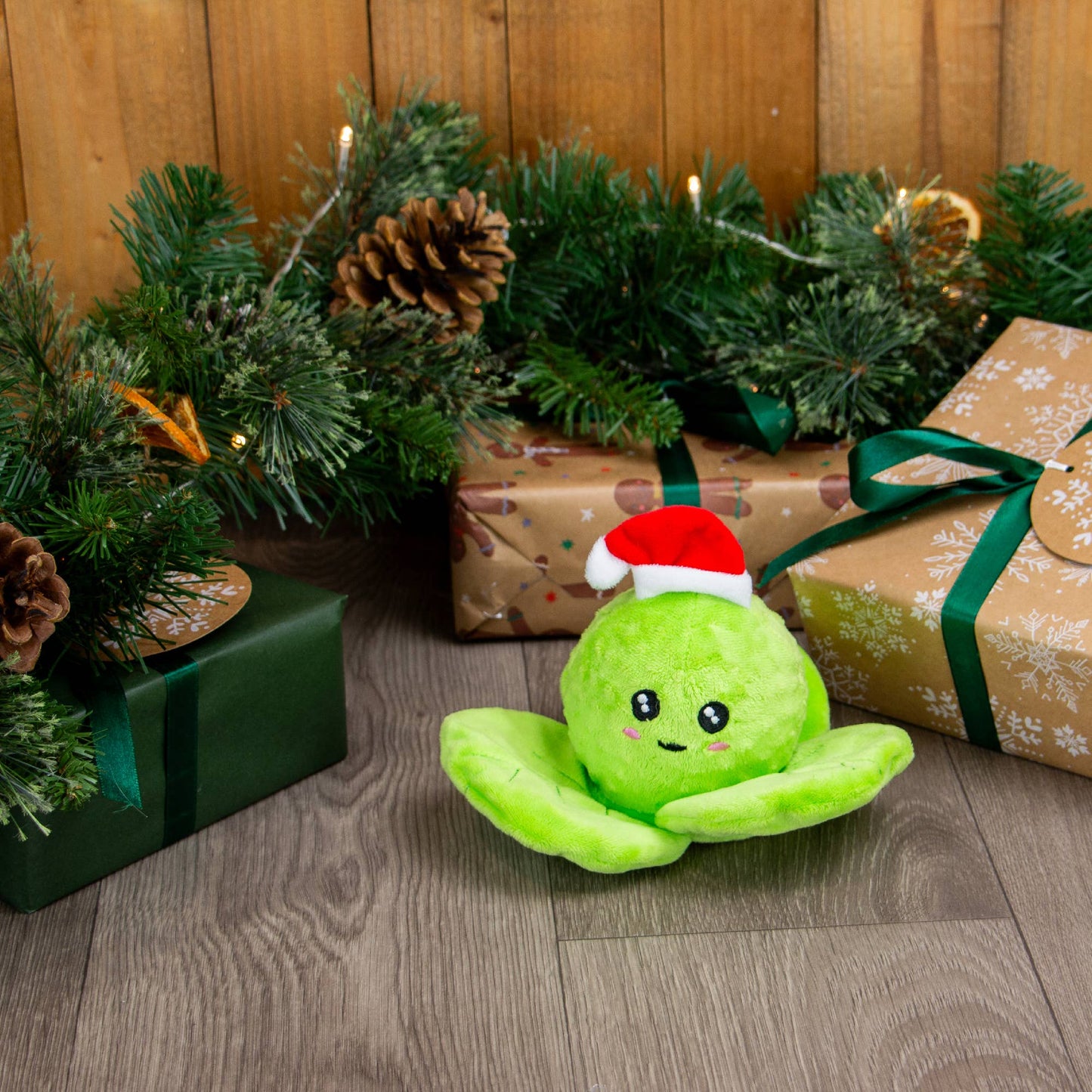 Green plush toy with a Santa hat on a wooden floor with Christmas decorations and presents.